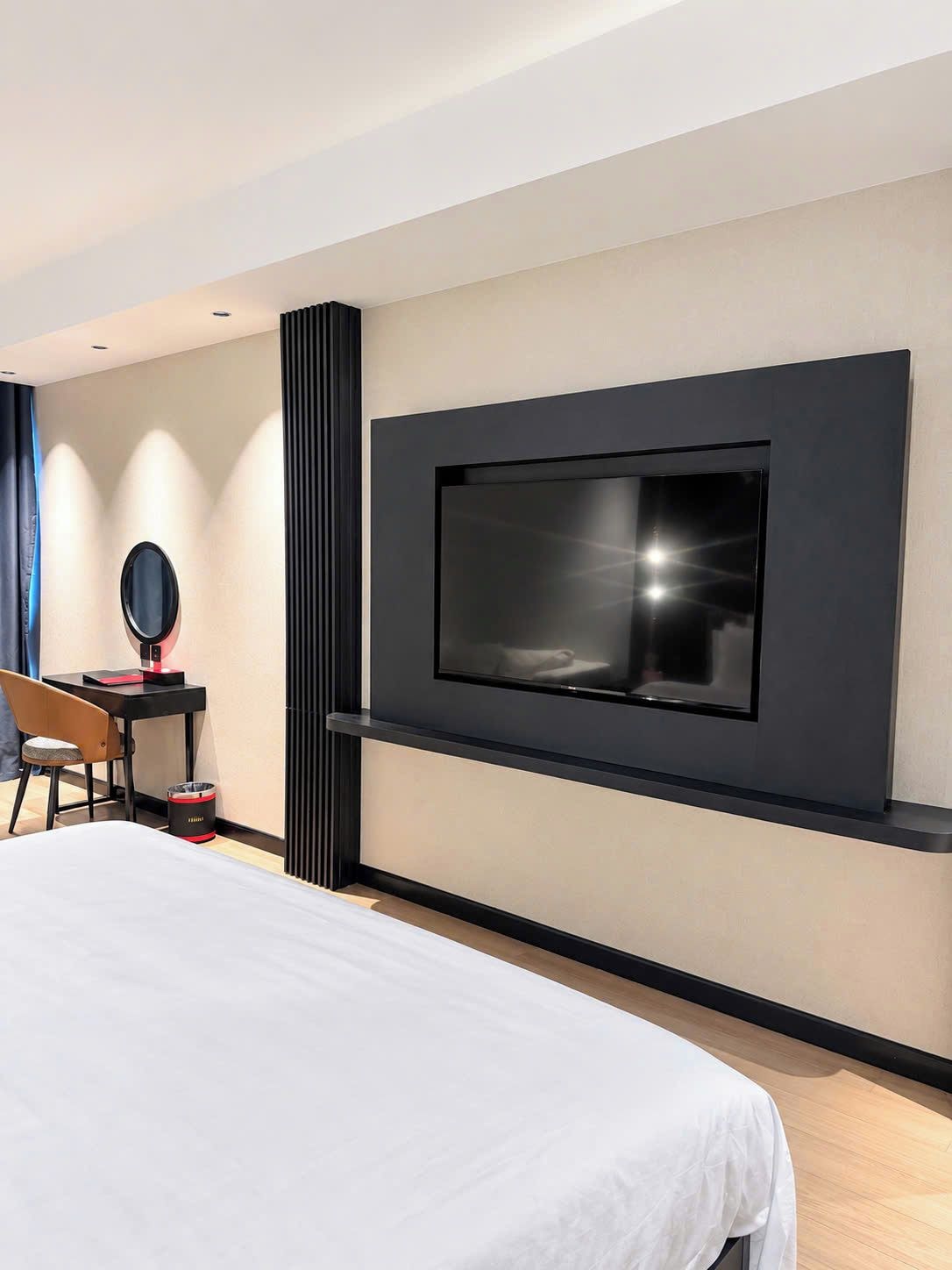 A modern hotel room with a large bed, a desk with a chair, a television mounted on the wall, and a trash bin.