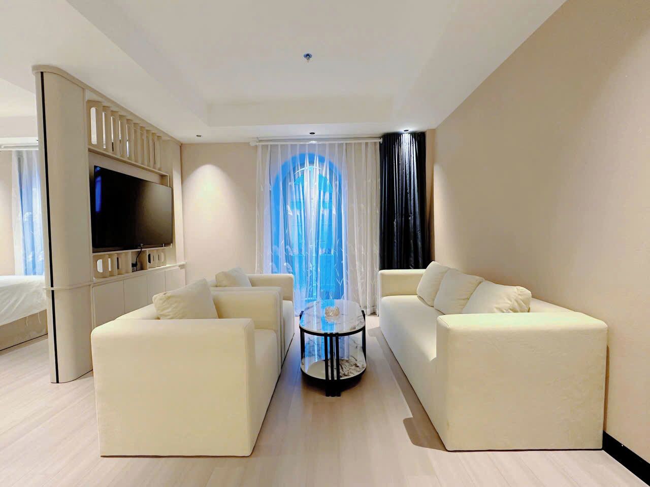 A modern living room with a flat-screen TV, two white sofas, a glass coffee table, and a large window with sheer curtains.
