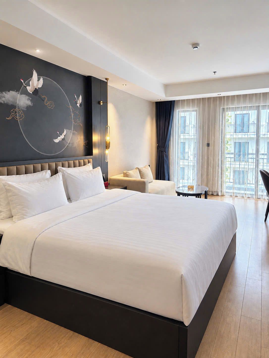A hotel room with a large bed, a sofa, a small table, and a view of the city through the windows.