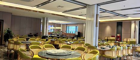 A spacious hotel dining area with round tables and wicker chairs, large windows, and a modern ceiling design.