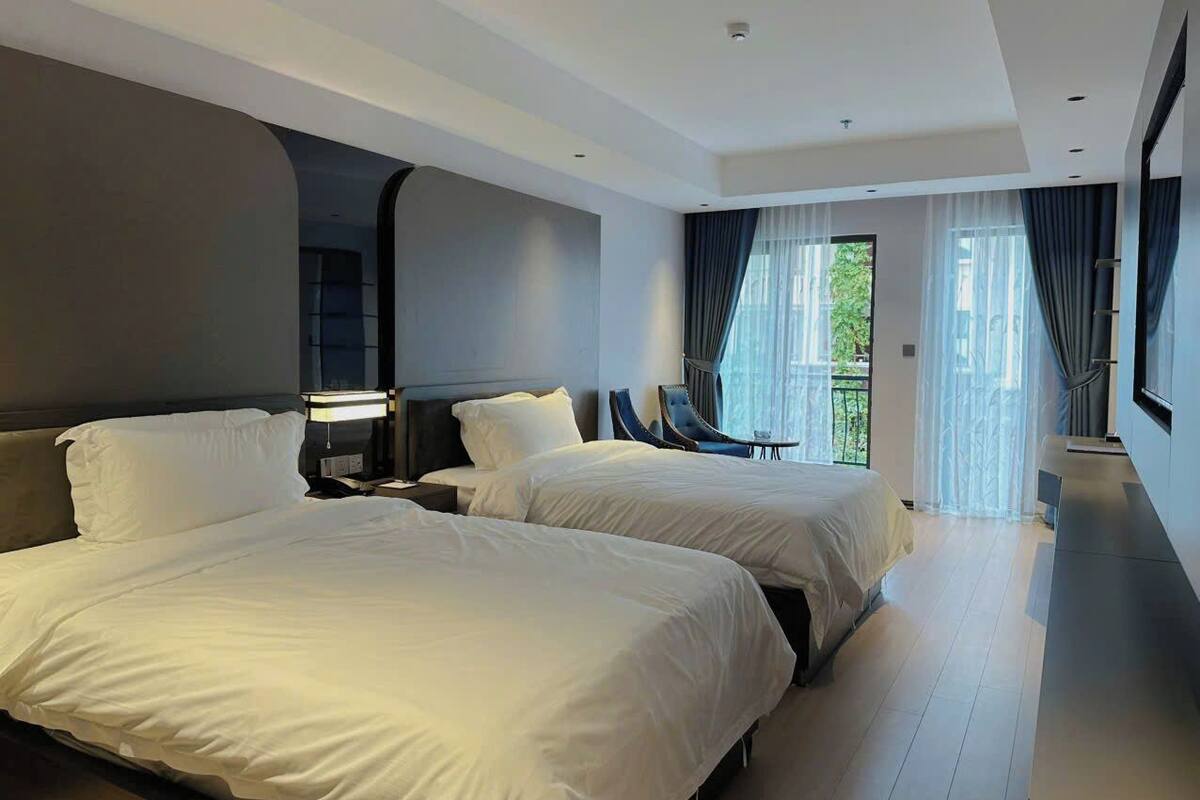 A modern hotel room with two beds, a large window with curtains, and a balcony.