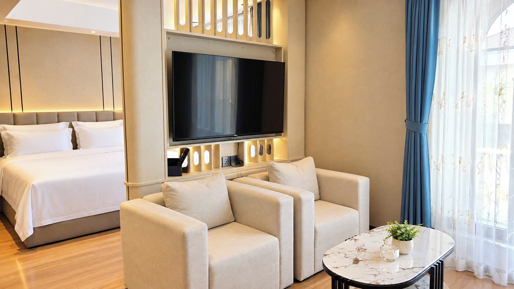 Habitación de hotel con cama, televisor montado en la pared, zona de estar con sofás y una mesita con planta.