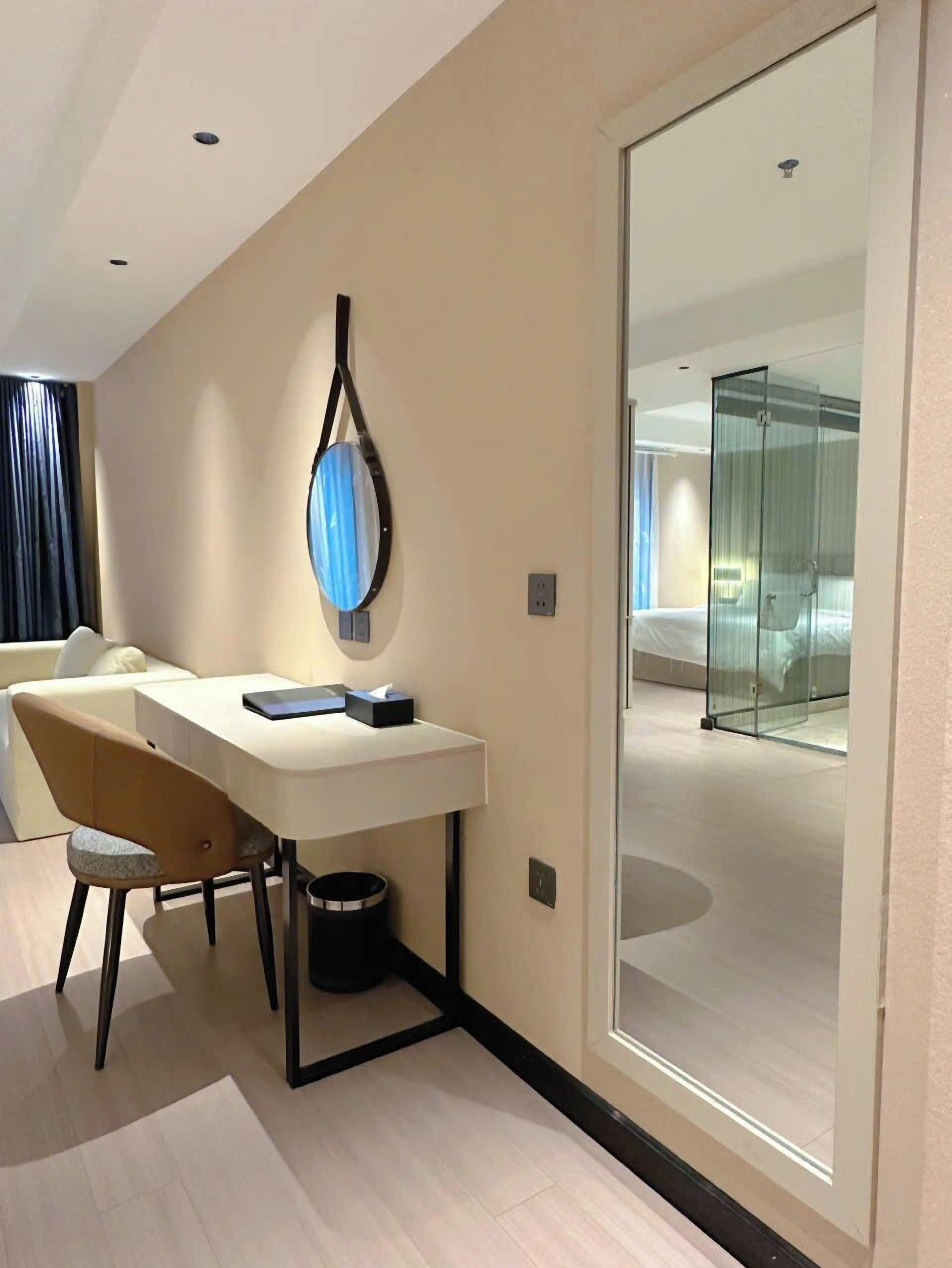 A modern hotel room with a desk, chair, and a glass partition separating the sleeping area.