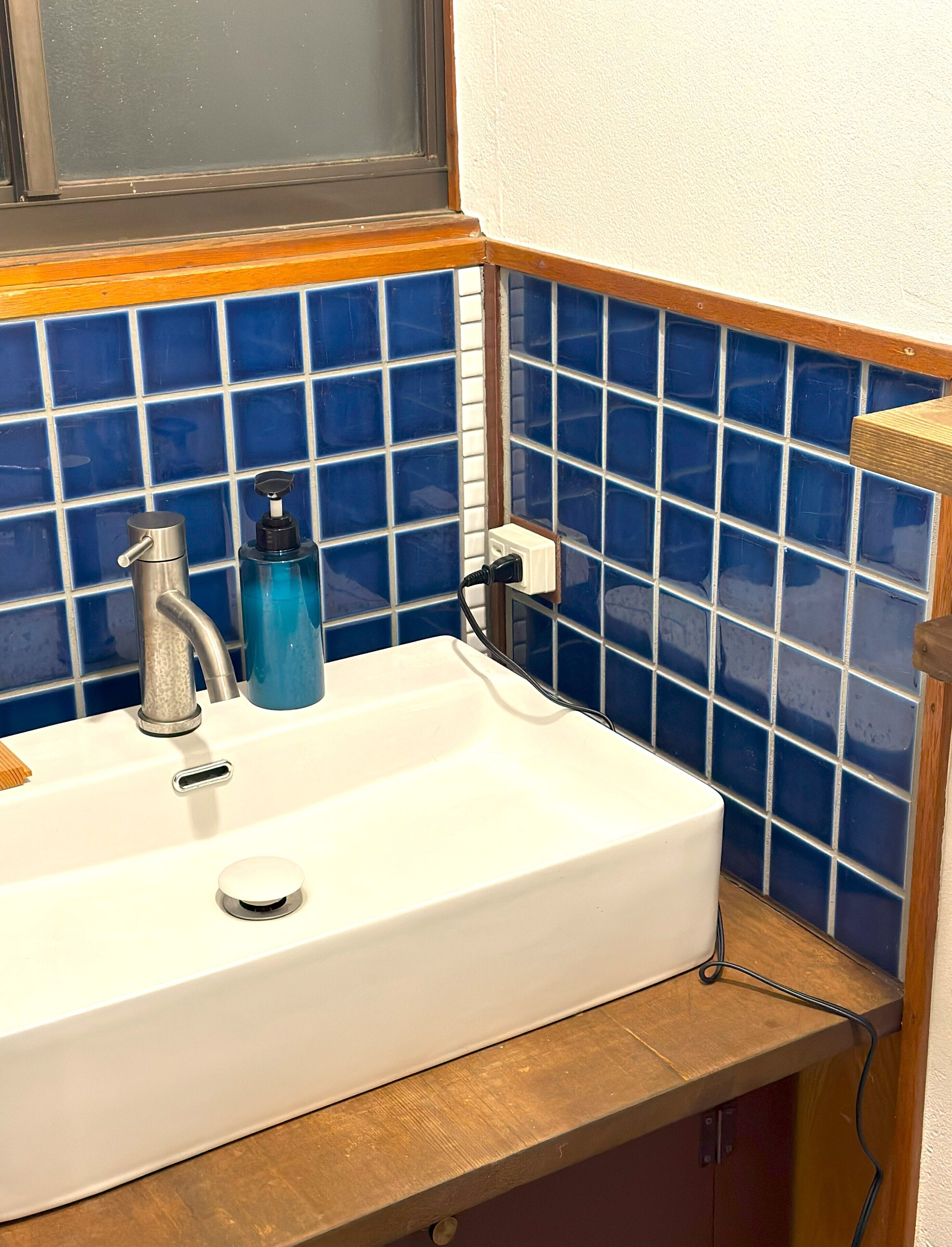 A white sink with a silver faucet, a blue soap dispenser, and a power outlet on a wooden countertop.