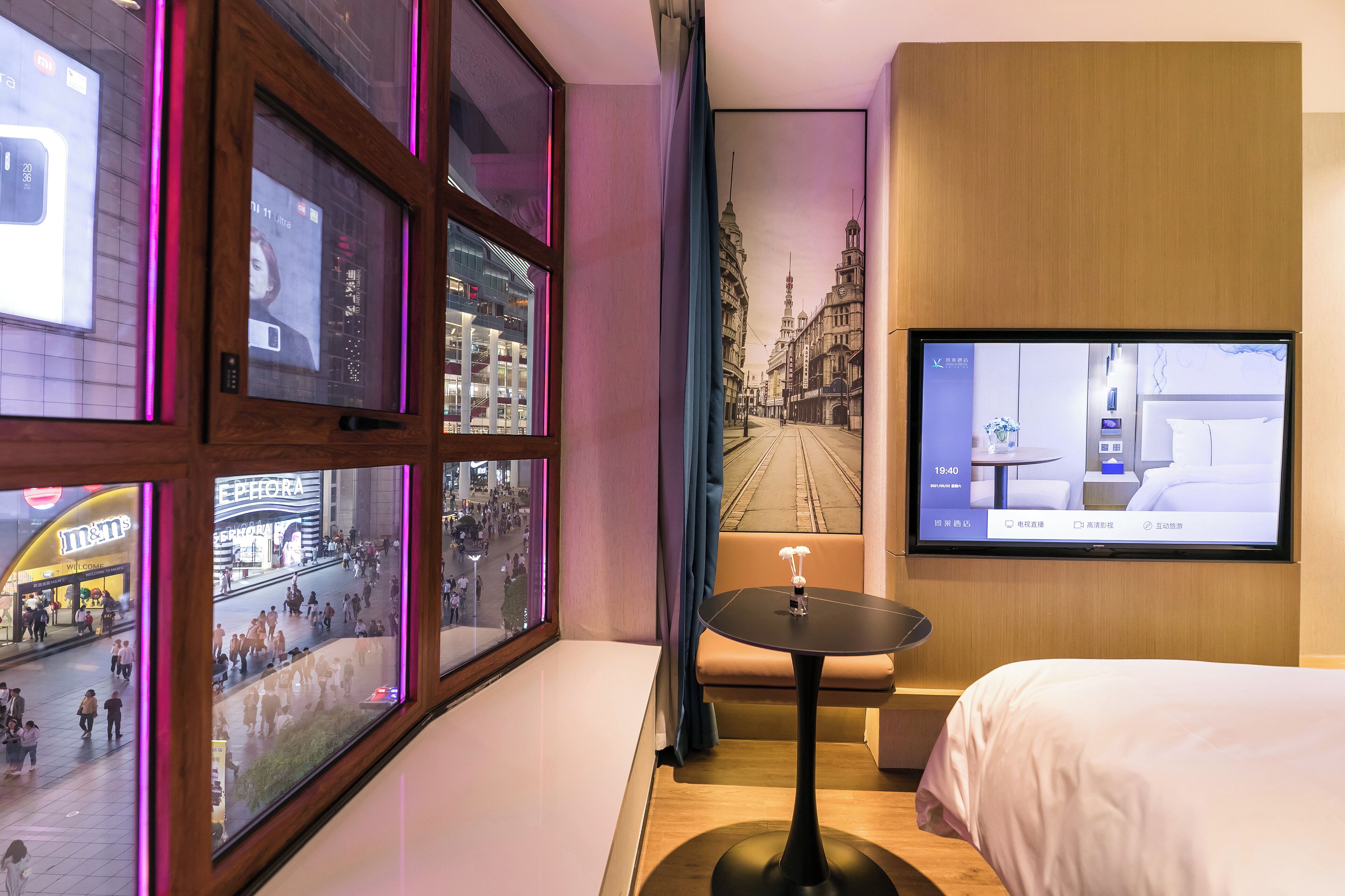 A hotel room with a large window, a bed, a small table, and a TV displaying a cityscape.