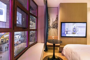 Een hotelkamer met een groot raam, een bed, een klein tafeltje en een televisie waarop een stadszicht te zien is.