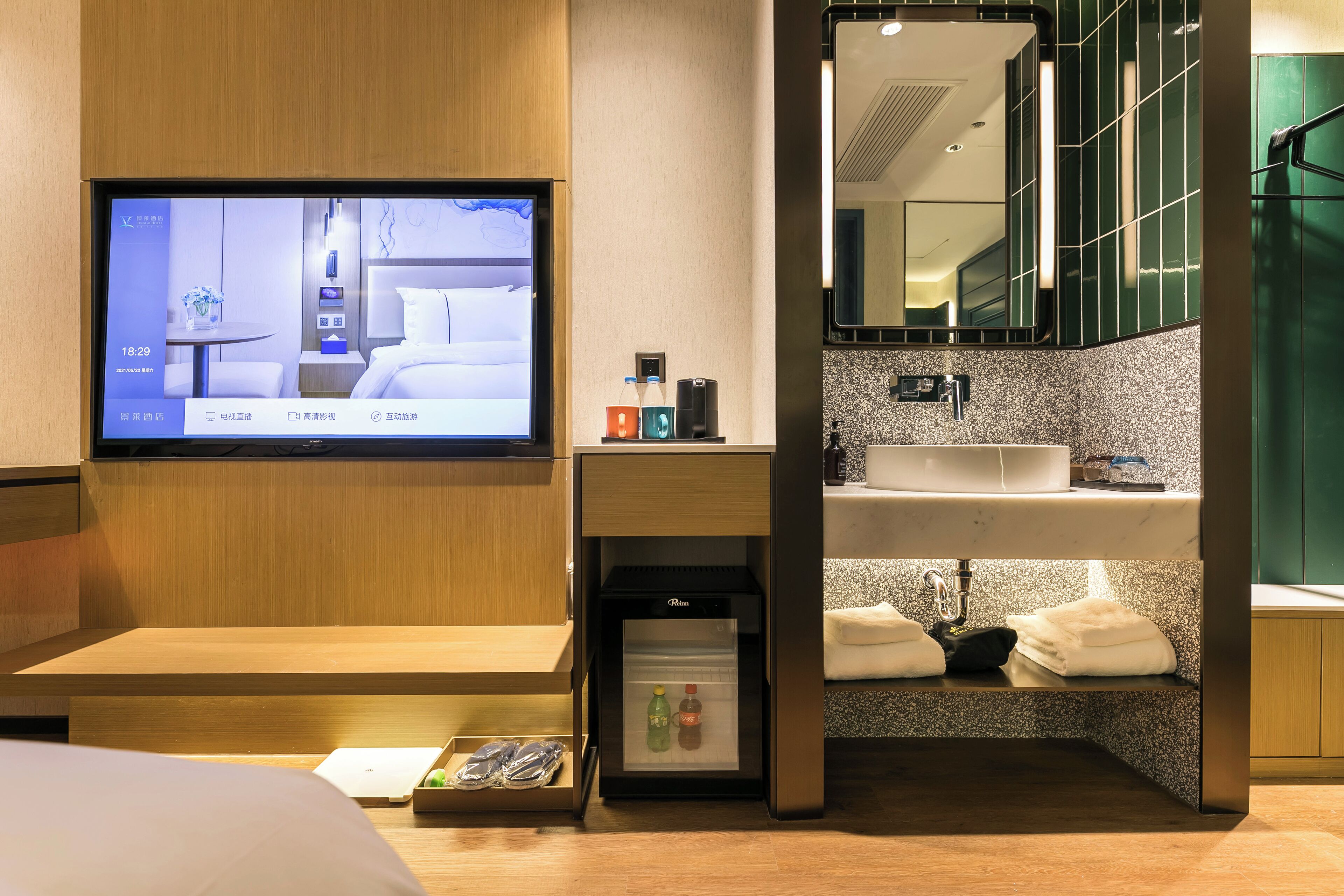 A modern hotel room with a television displaying a room layout, a minibar, a sink with towels, and a mirror above the sink.