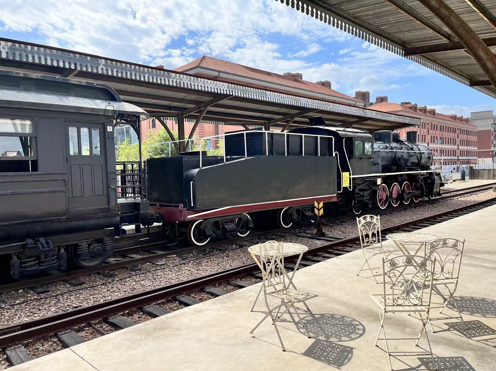A train station with two locomotives on the tracks, a covered waiting area with metal chairs, and a building with a red roof in the background.