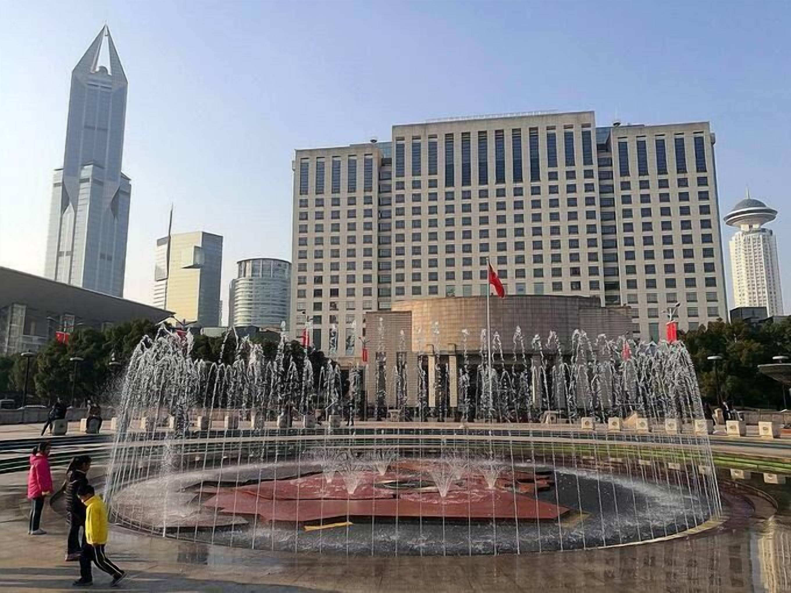 Un'ampia fontana pubblica con numerosi getti d'acqua disposti in un design circolare, circondata da un'area lastricata dove passeggiano alcune persone. Sullo sfondo, si vedono diversi edifici alti, tra cui un moderno grattacielo.