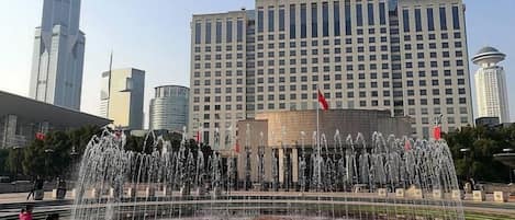 Una gran fuente pública con múltiples chorros de agua en un diseño circular, rodeada de una zona pavimentada con algunas personas caminando. Al fondo, varios edificios altos, incluyendo un rascacielos destacado.