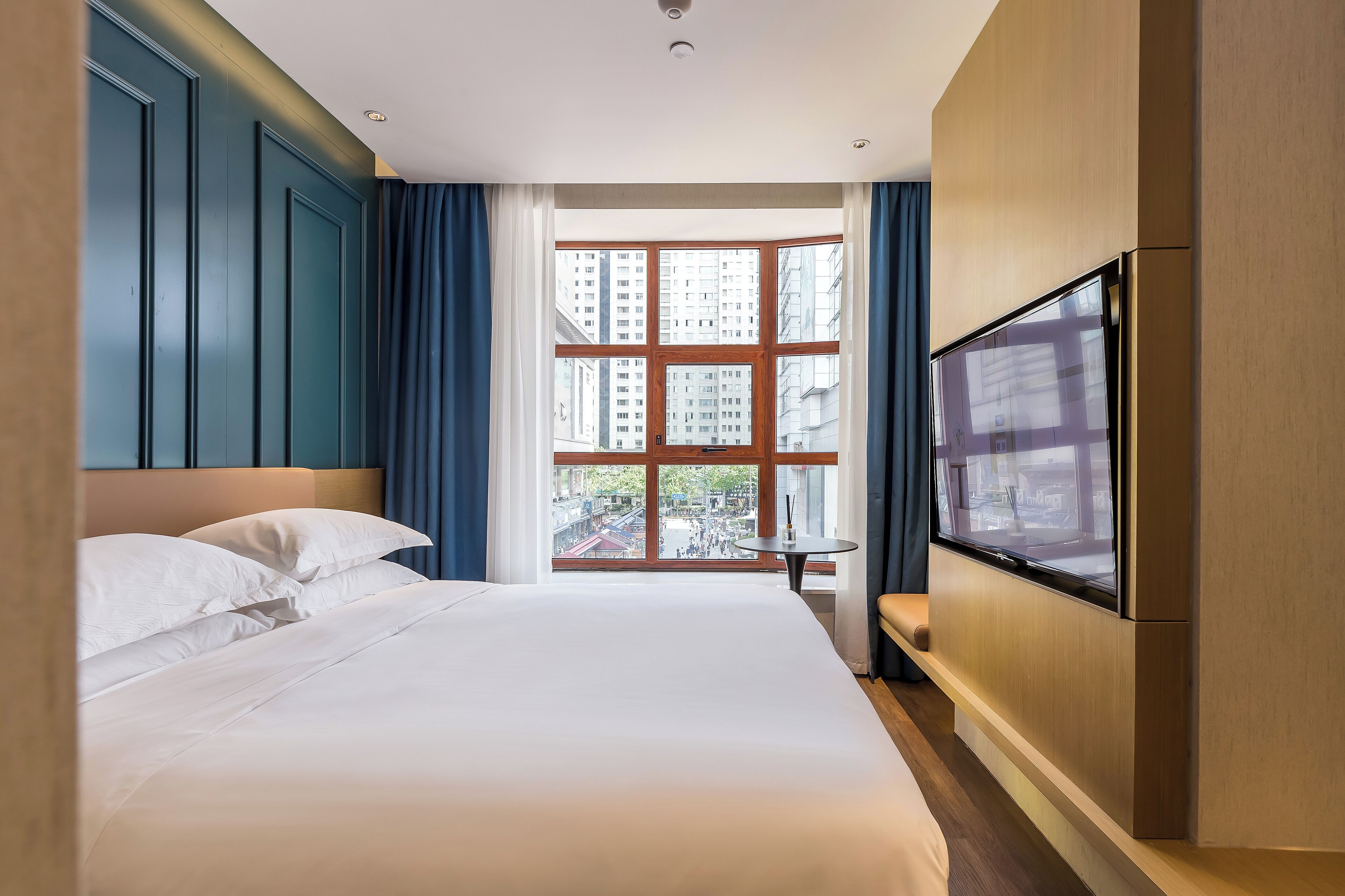 A hotel room with a large bed, a TV mounted on the wall, and a window with a view of buildings and a courtyard.