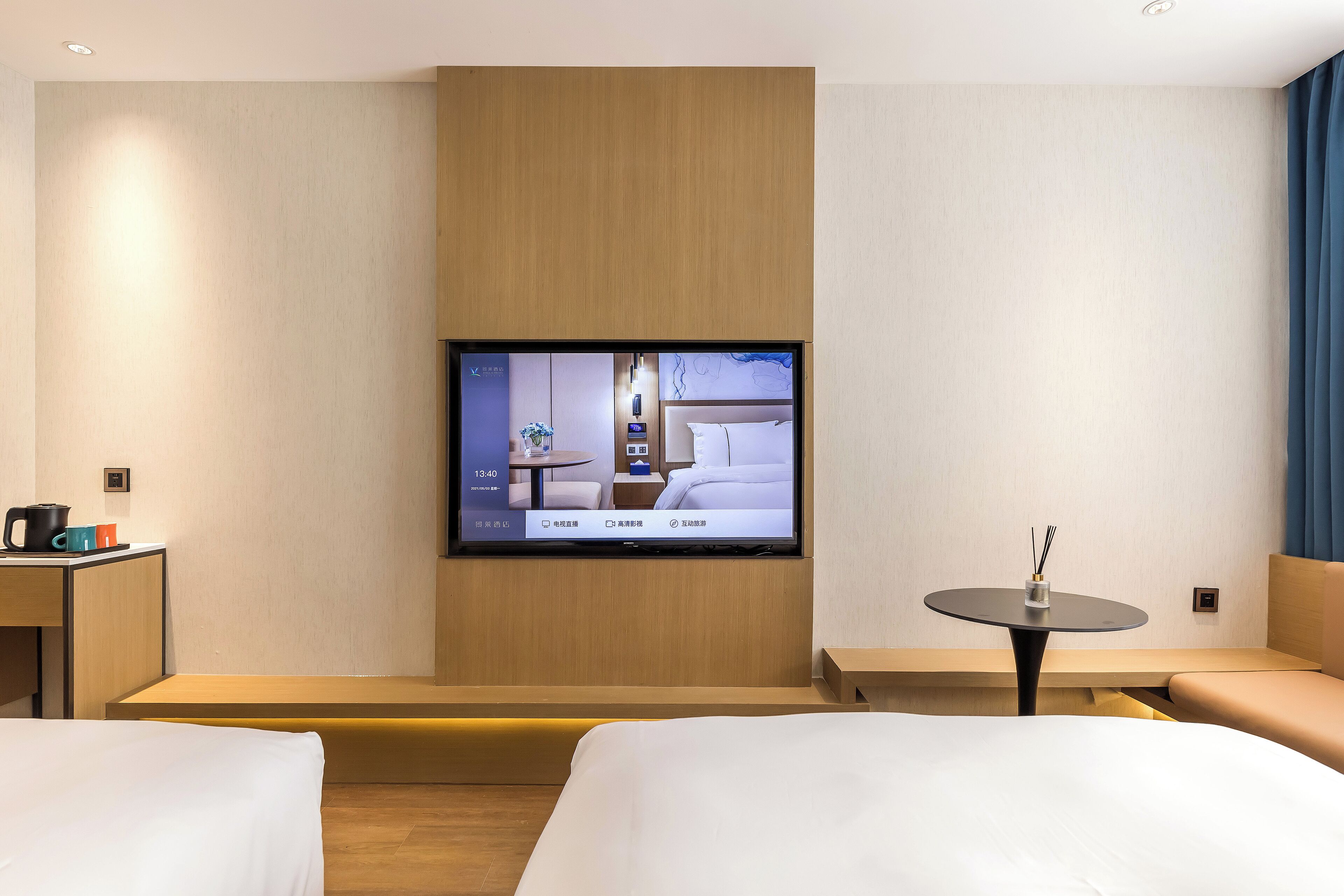 A modern hotel room with a large flat-screen TV mounted on the wall, a wooden bench, a small round table with a candle, and a bed with a white comforter.