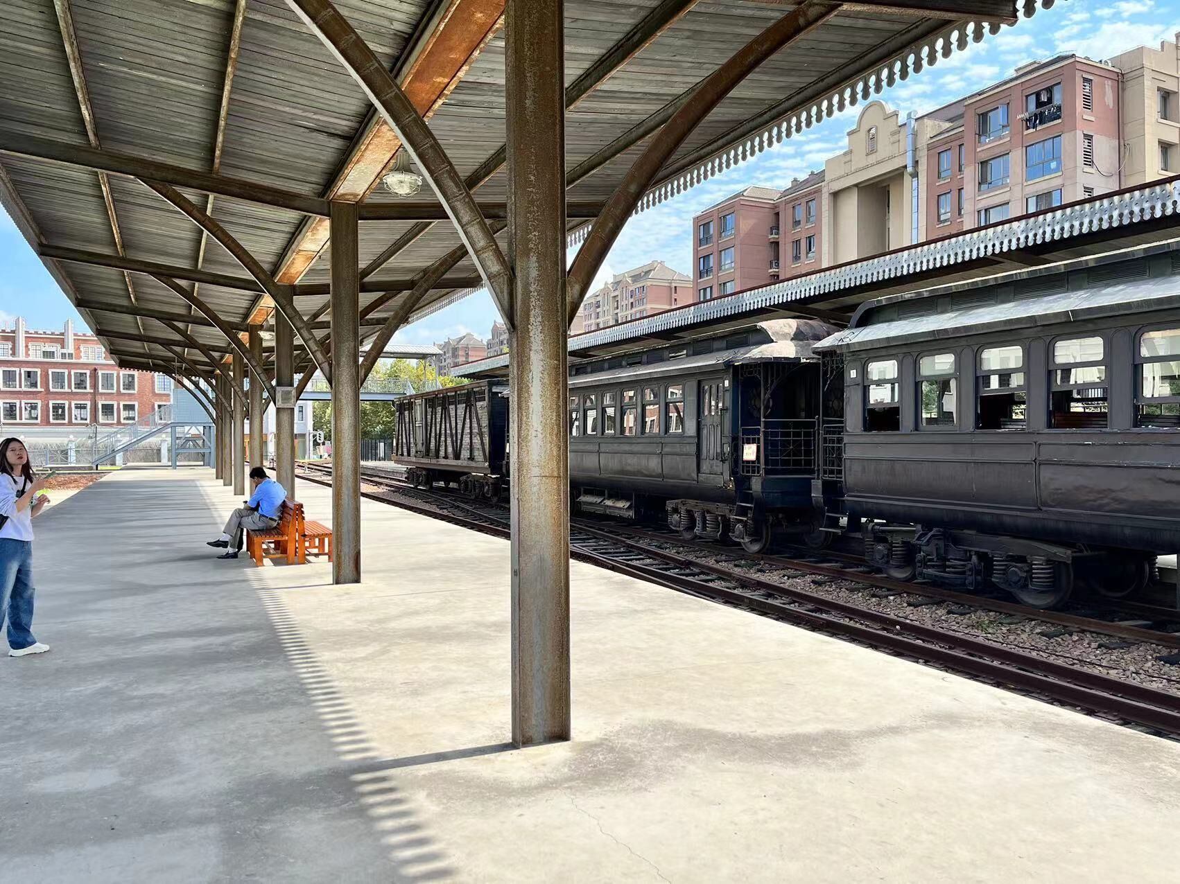 Una banchina di stazione ferroviaria con un'area di attesa coperta, panchine e un treno sui binari.