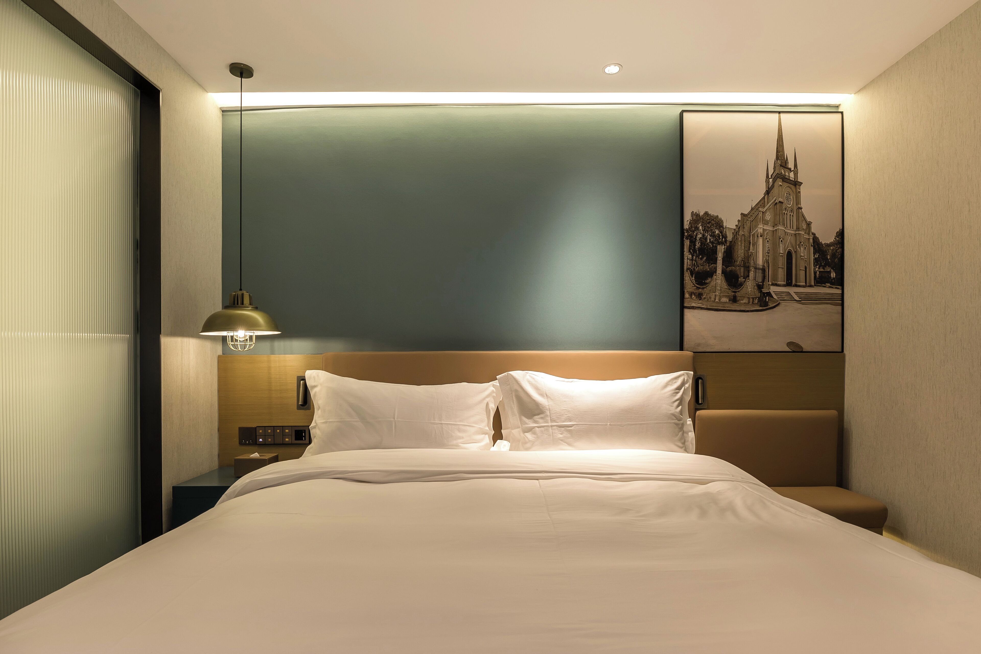 Een hotelkamer met een bed, een nachtkastje, een lamp en een aan de muur hangend schilderij van een kerk.