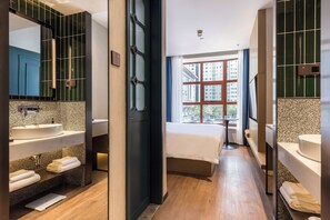 A modern hotel bathroom with two sinks, a large mirror, and a bed with a view of buildings through the window. - Jinglai Hotel (Shanghai Bund East Nanjing Road Pedestrian Street) (Shanghai)