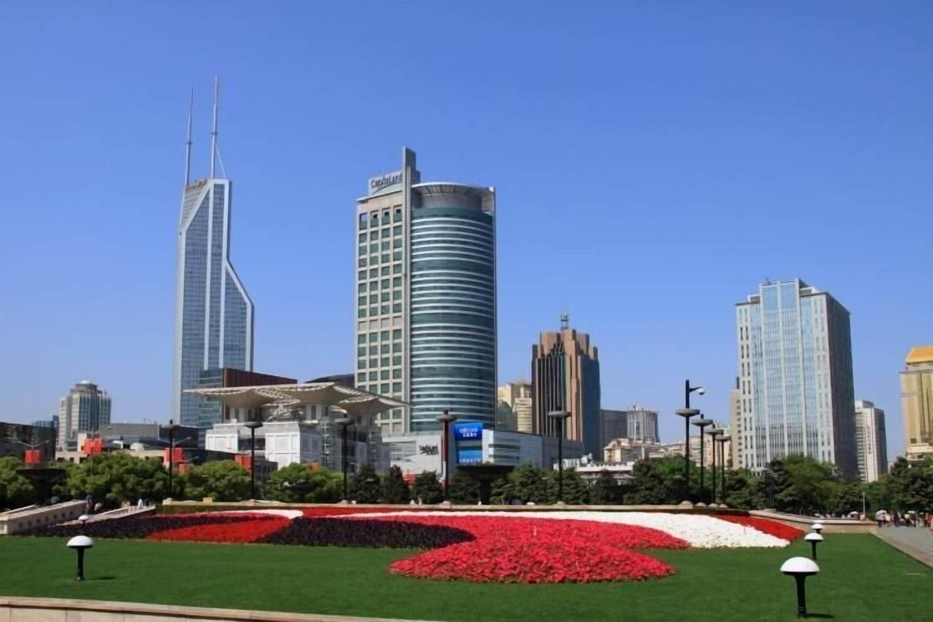 Paisagem urbana com edifícios modernos e altos, um grande jardim de flores e um céu azul claro.