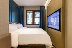 A hotel room with a bed, a TV mounted on the wall, a window with curtains, and a view of buildings outside.