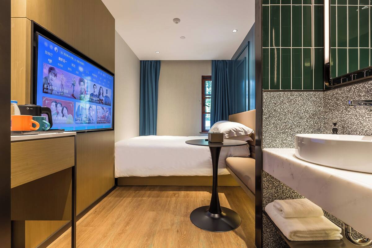 Une chambre d’hôtel moderne, équipée d’un lit, d’une petite table ronde, d’une télévision et d’une salle de bain avec un lavabo et un miroir, ainsi que d’un plancher en bois.