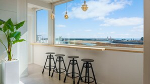 Habitació amb un gran finestral que dona a una platja, una planta en test i quatre tamborets.
