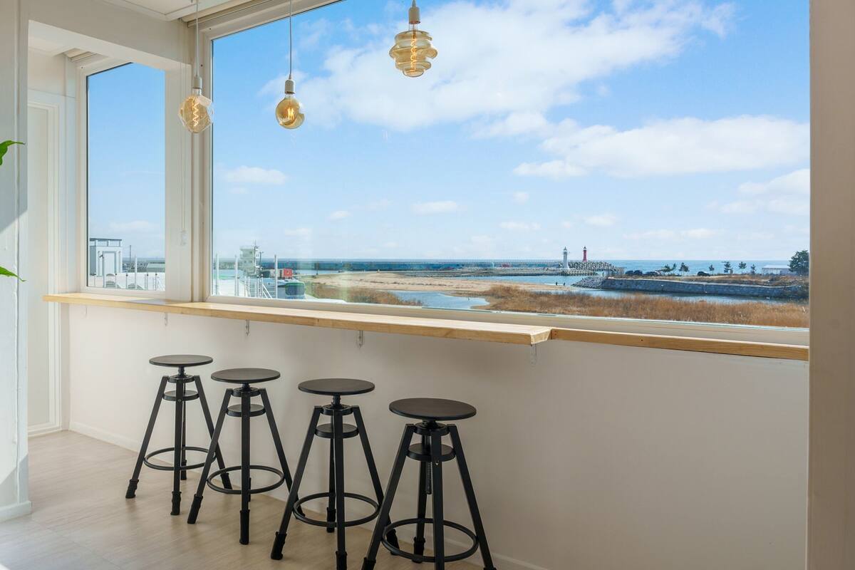 Ein Zimmer mit großem Fenster und Blick auf einen Strand, einer Topfpflanze und vier Barhocker.