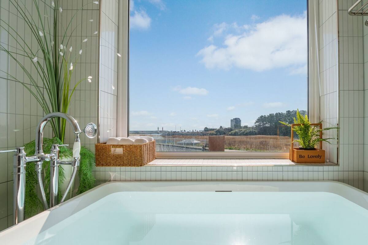 Ein modernes Badezimmer mit einer großzügigen Badewanne, einem Fenster mit Blick auf die Stadt und einer Topfpflanze.