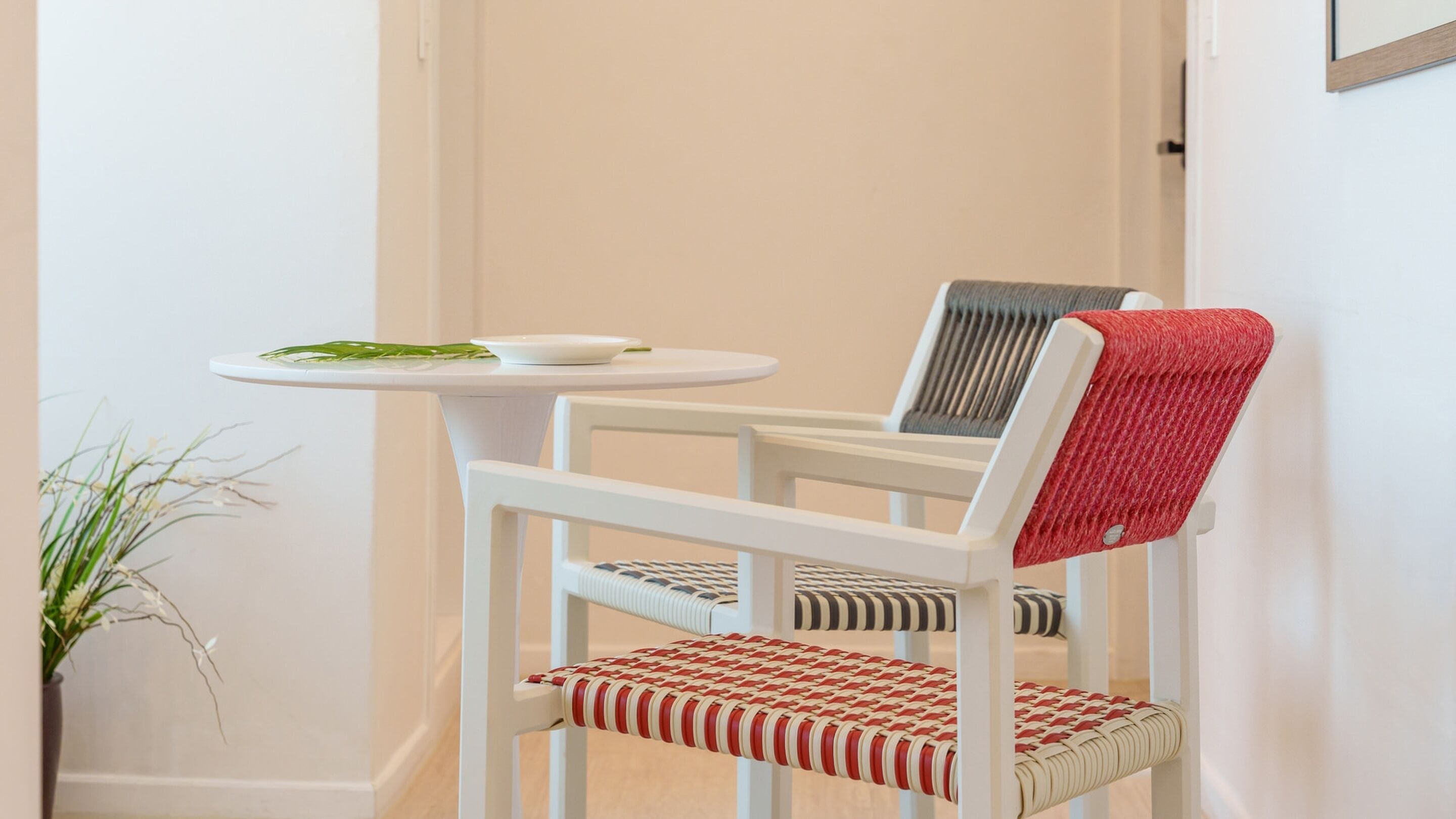 A white chair with a red and white woven seat and backrest, a small white table with a white plate and green leaves, and a potted plant to the left.