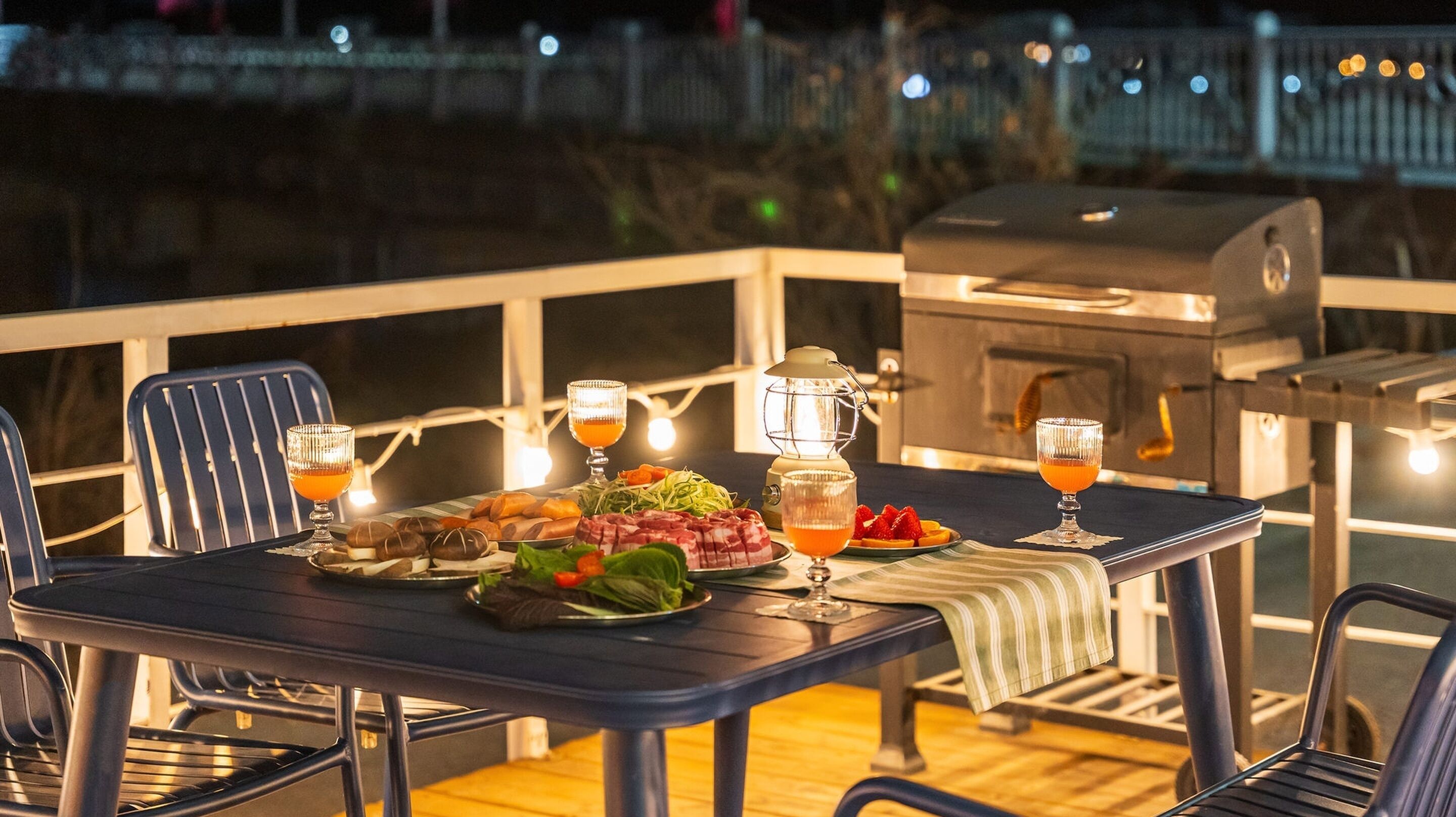 An outdoor dining area with a grill, string lights, and a table set with food and drinks.