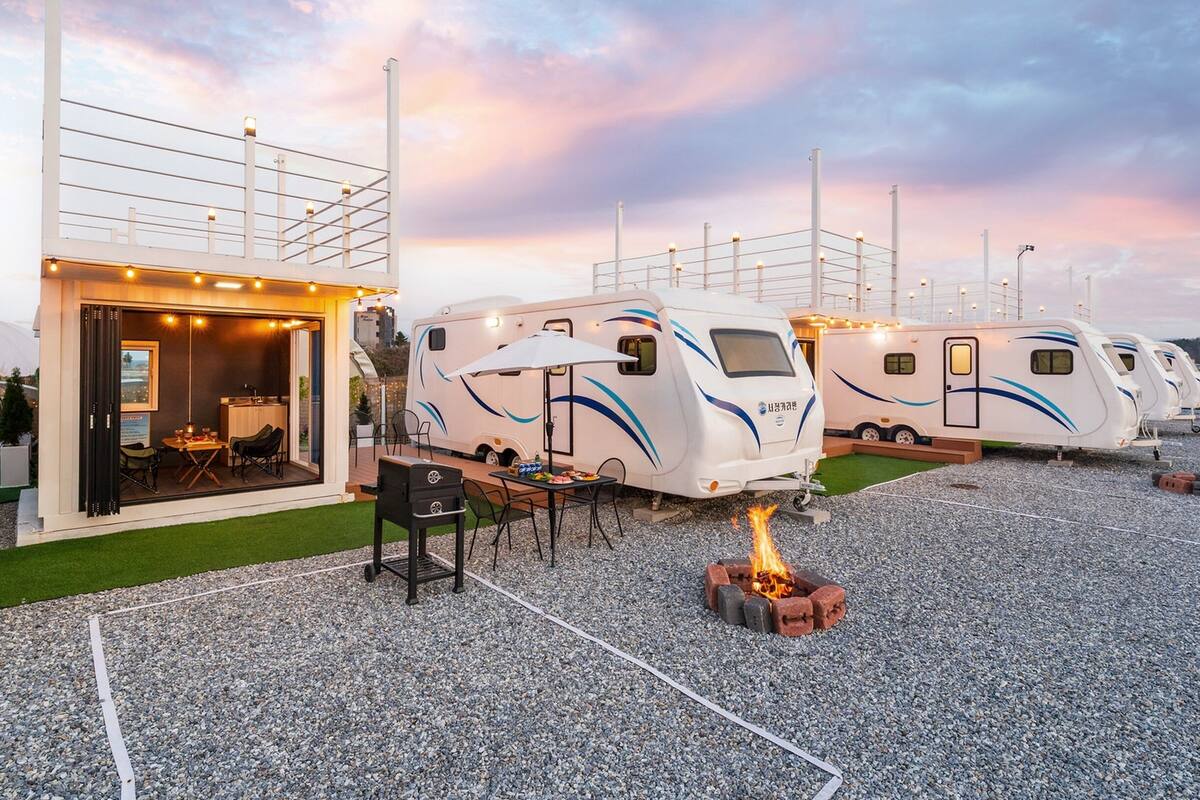 Une rangée de camping-cars est stationnée sur une surface de gravier, à proximité d’un bâtiment doté d’une porte en verre et d’une terrasse.