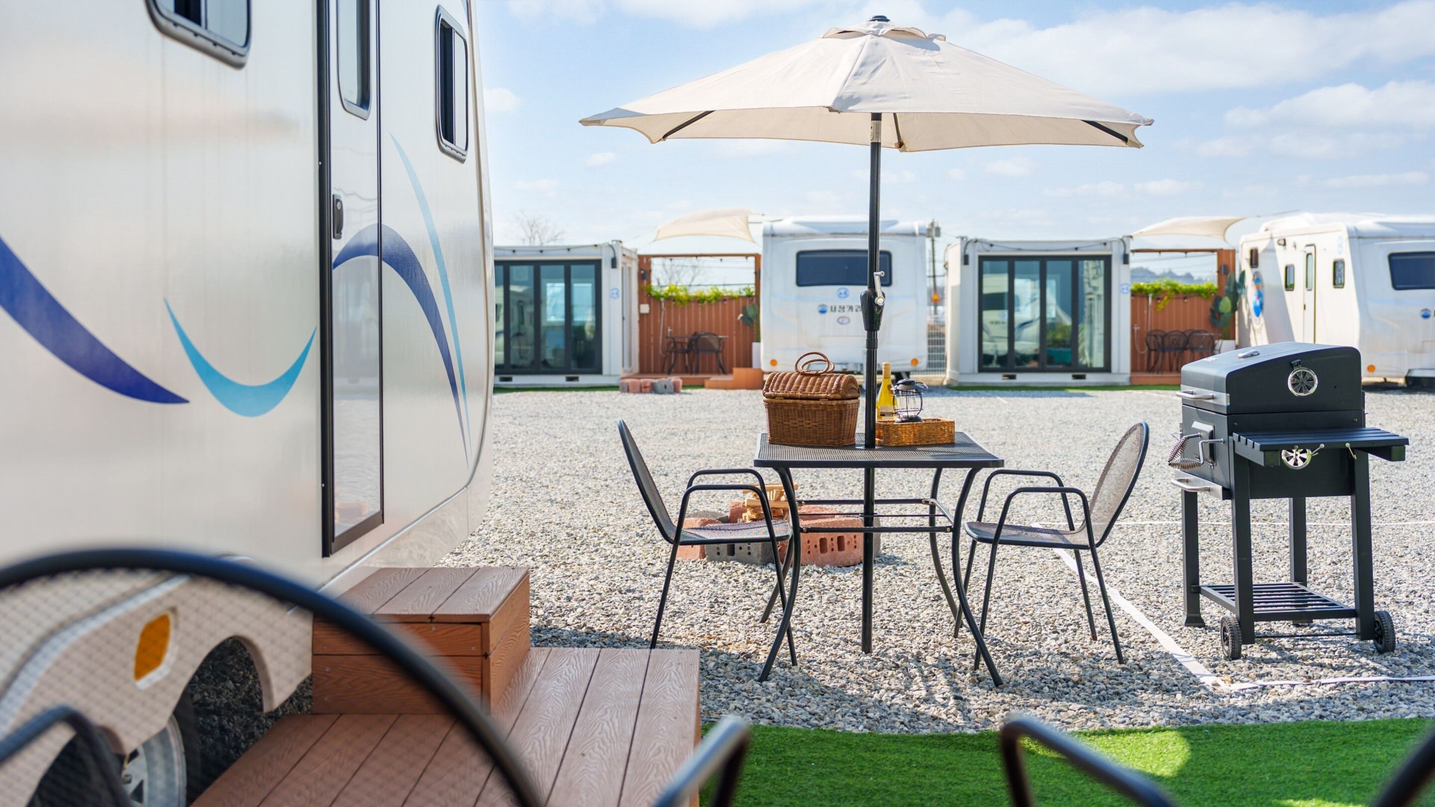 A campsite with a table and chairs under an umbrella, a barbecue grill, and RVs in the background.