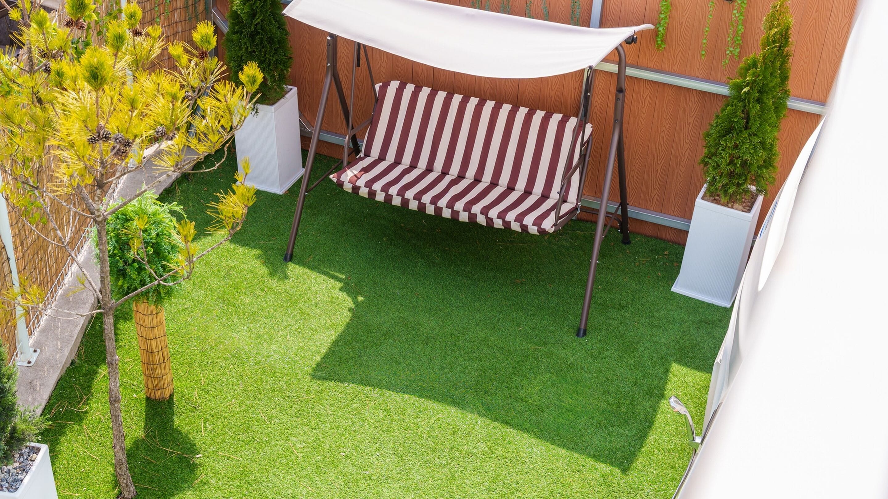 A patio with a striped swing, artificial grass, and potted plants.
