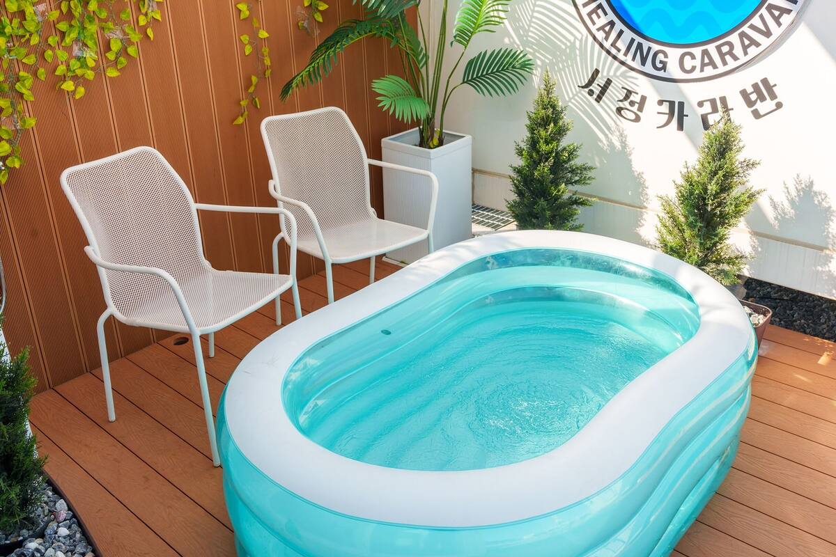 Une piscine extérieure dotée d’un bord gonflable blanc, entourée d’une terrasse en bois. On y trouve deux chaises en métal blanc et un pot de fleurs sur la gauche.