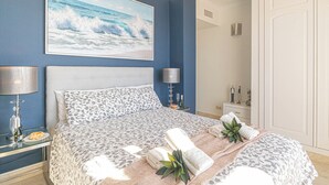 Een slaapkamer met een groot bed, twee nachtkastjes met lampen, een bijzettafeltje met een fles en glazen, en een schilderij van de oceaan aan de muur.