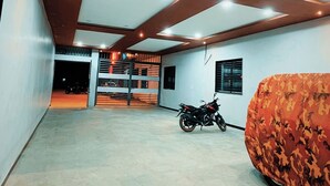 Et rummeligt indendørs område med en motorcykel parkeret ved en væg, en seng med camouflagedug og en trappe til en etage over.