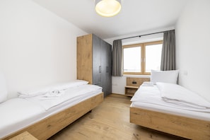 Ein moderner, gut beleuchteter Raum mit zwei Holzbettgestellen, einem grauen Kleiderschrank und einem Fenster mit Vorhängen.