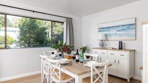 A dining area with a wooden table set for four, white chairs, a white cabinet with drawers, and a large window with curtains. - Bangalee 2 at Sunrise - waterfront apartment (Soldiers Point)