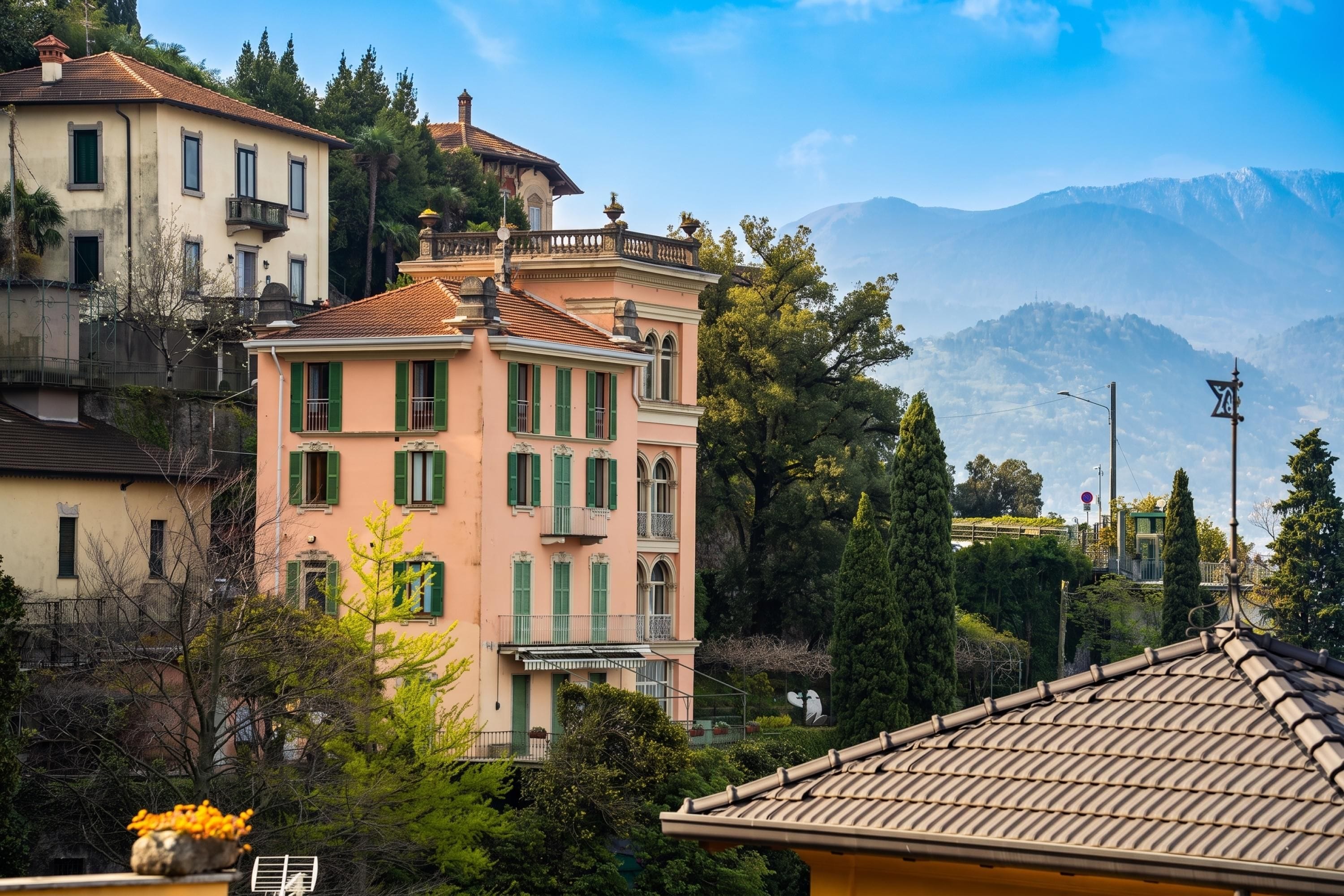 A multi-story building with a balcony, surrounded by greenery and mountains in the background.