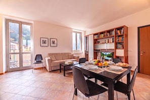 Uma sala de jantar com mesa posta para quatro pessoas, um sofá, uma estante de livros e uma televisão.