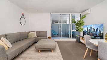A modern living room with a grey sofa, a grey ottoman, a dining table with chairs, a large TV, and a sliding glass door leading to a balcony with chairs.