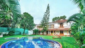 A swimming pool with a tiled edge, surrounded by lush greenery and a house with a tiled roof. - Peacock House Saligao (Saligao)