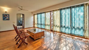A spacious room with a wooden floor, a ceiling fan, a wooden coffee table, and a large window with patterned curtains. - Peacock House Saligao (Saligao)