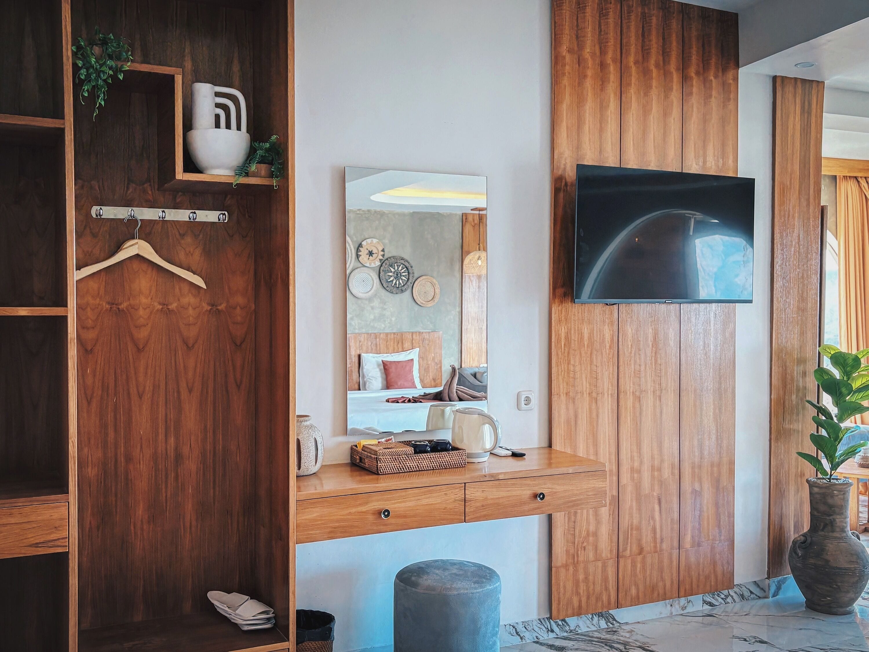 A modern bedroom with a wooden wardrobe, a bedside table with a mirror, a TV mounted on the wall, and a potted plant.