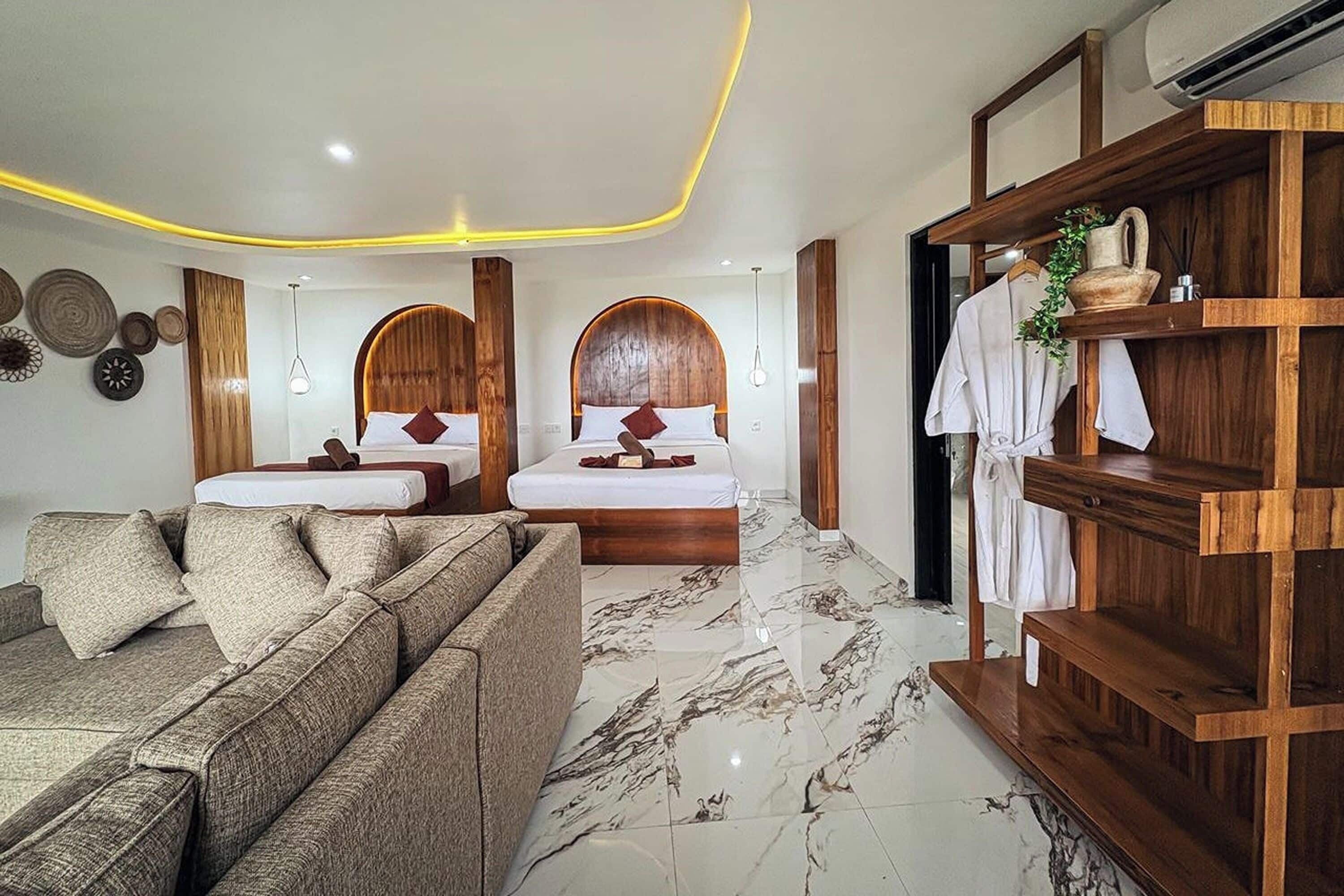 A modern hotel room with two beds, a sofa, a wooden shelf with towels, and decorative wall art.