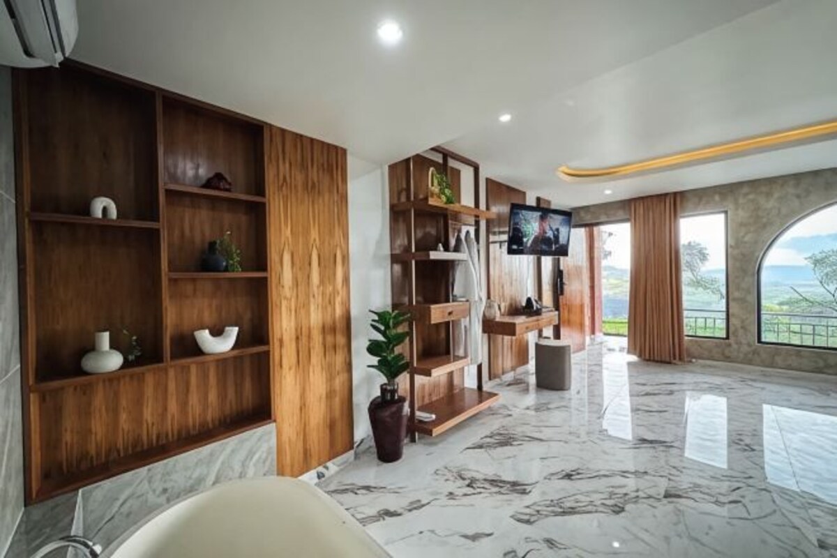 A modern bathroom with a bathtub, wooden shelving, a flat-screen TV, and a large window with a view.