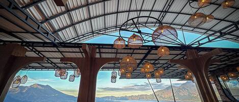 Terraza con vistas a montañas y una masa de agua, con lámparas colgantes y mobiliario de madera.