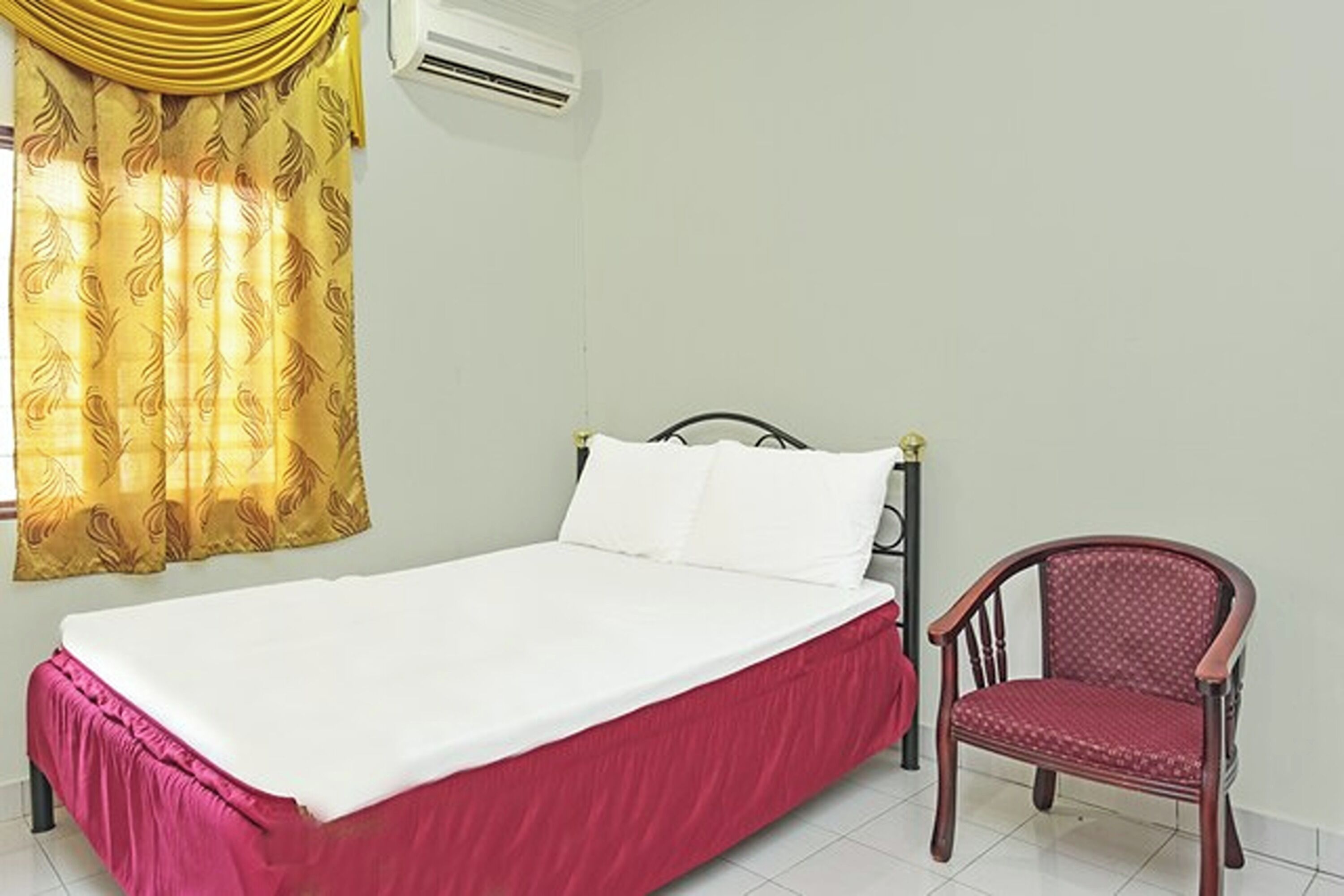 Un lit simple avec une couvre-lit rouge et des oreillers blancs, une chaise en bois et un rideau jaune à motifs.