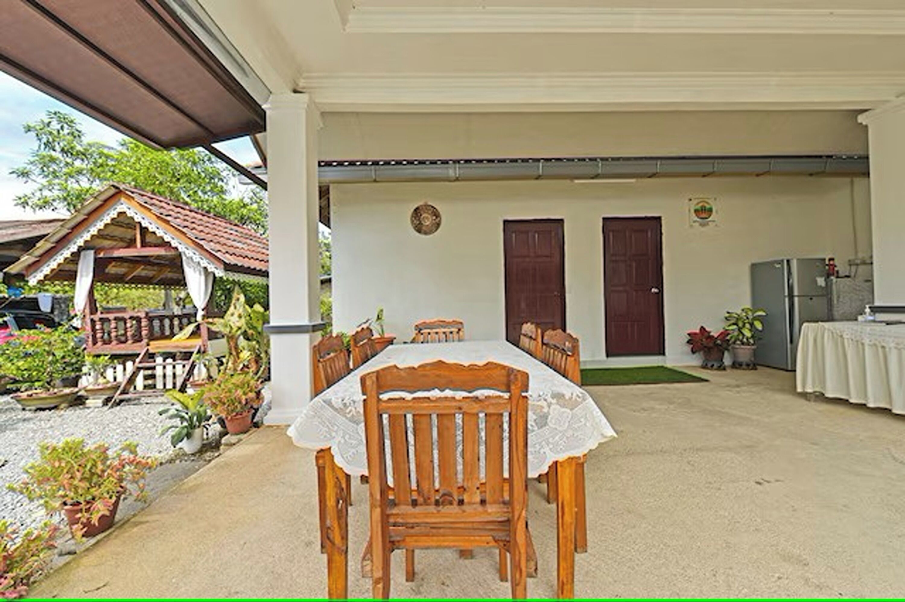 A covered outdoor dining area with a wooden table and chairs, surrounded by potted plants and a small wooden structure.