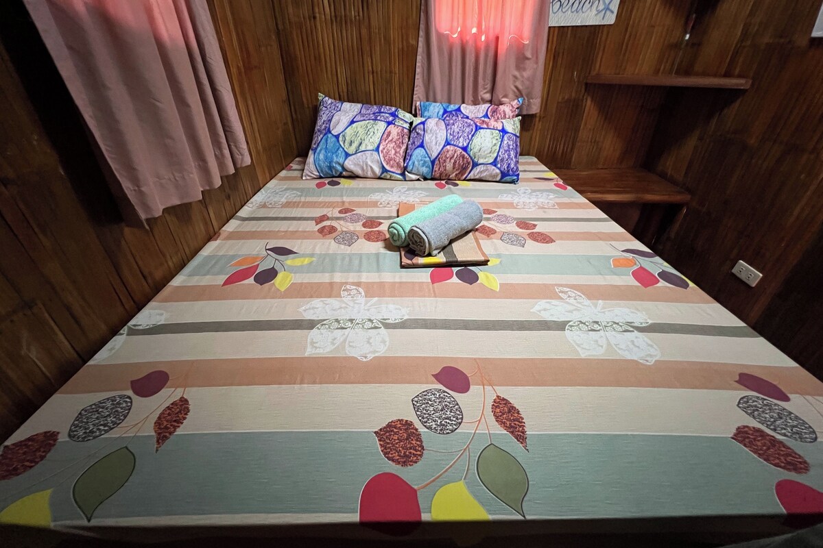 Un lit recouvert d’une courtepointe aux motifs de feuilles colorées, deux oreillers, une serviette enroulée et un panneau accroché au mur.