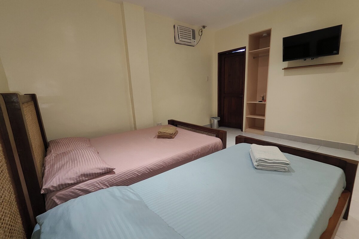 A room with two beds, one with a pink bedspread and the other with a blue bedspread, both with folded towels on them. There is a TV mounted on the wall above the beds.