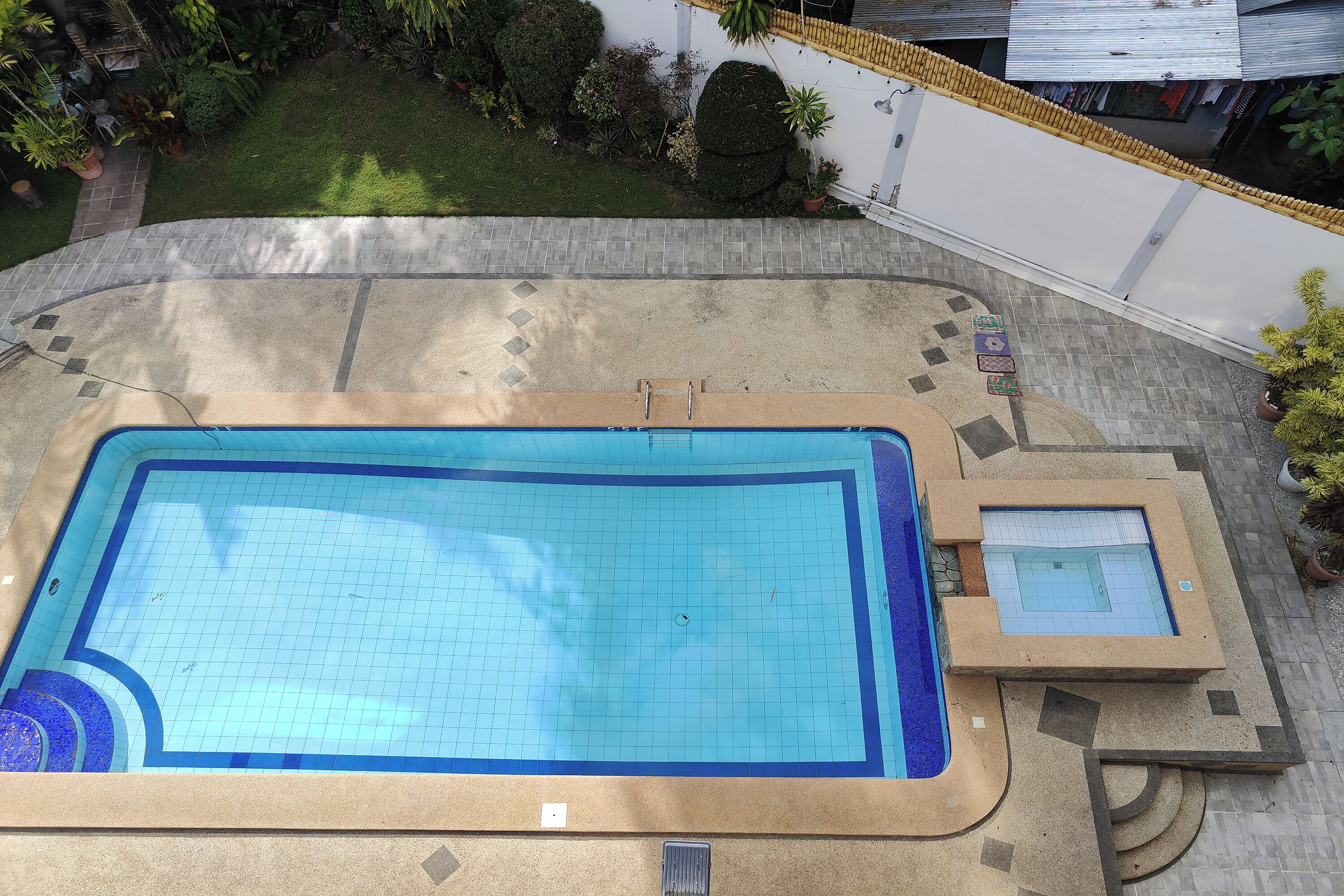 Et rektangulært swimmingpool med flisebelagte omgivelser, et mindre tilstødende bassin og en flisebelagt sti, der fører til en hvid væg med gul kant.