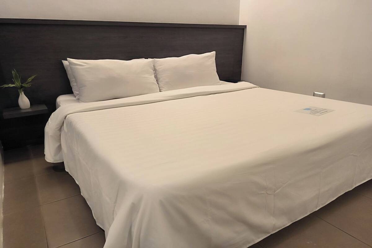 A neatly made bed with white bedding, a dark headboard, and a small bedside table with a white vase and a plant.